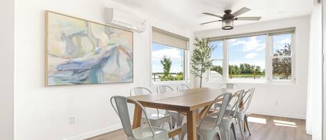 A dining area with a wooden table and light gray chairs, large windows with a view of greenery, and a ceiling fan.