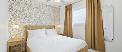 A bedroom with a bed, a ceiling fan, a nightstand, a mirror, and curtains.
