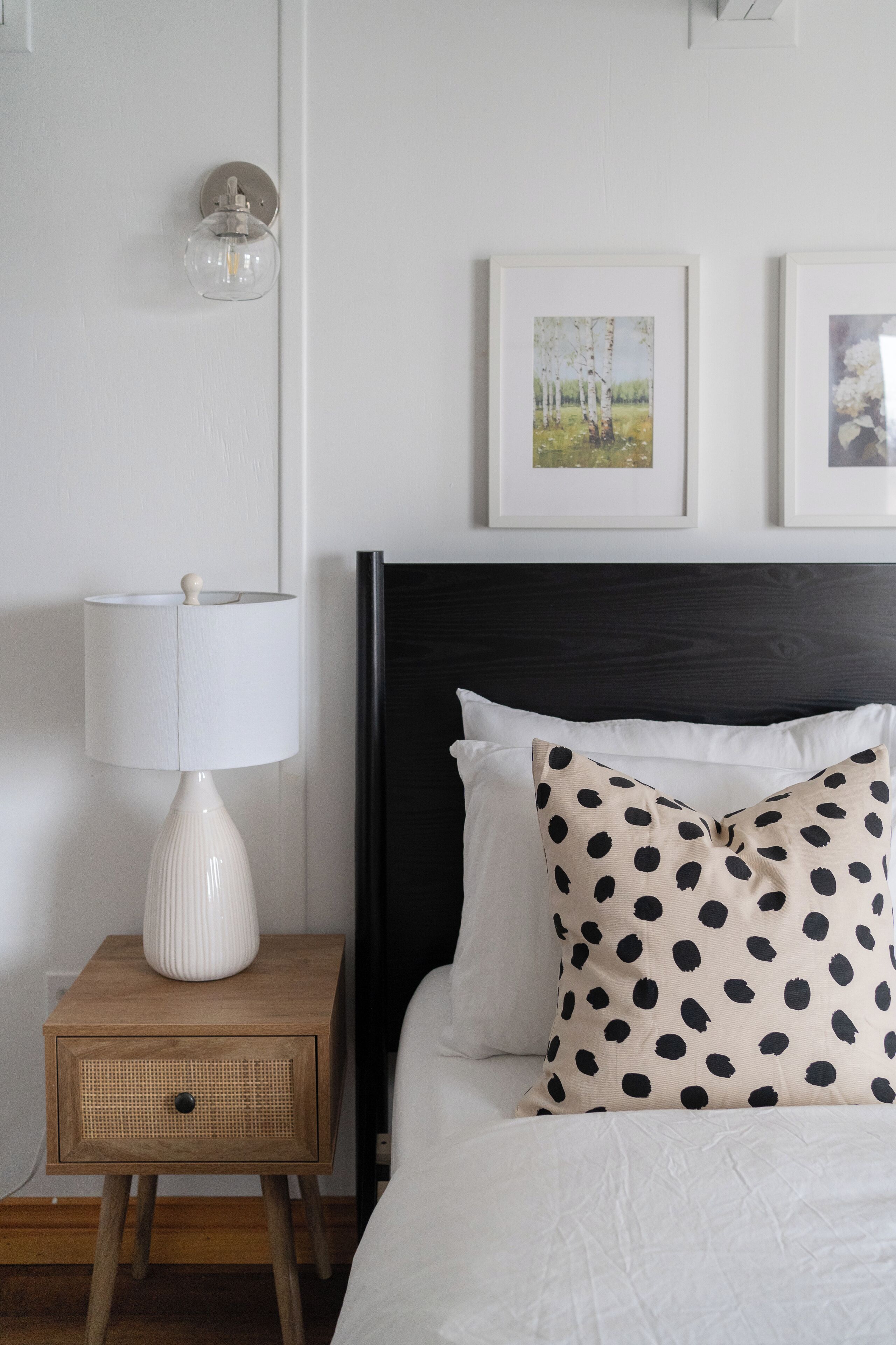 Een keurig opgemaakte slaapkamer met een bed, een bedlampje, een klein houten nachtkastje en twee ingelijste foto’s aan de muur.