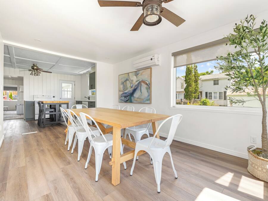 A spacious dining area with a wooden table and white chairs, a ceiling fan, and a view of the outside through large windows.