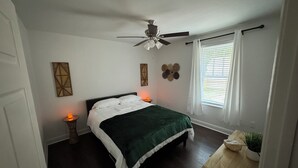 Ein Schlafzimmer mit Bett, Deckenventilator, Nachttisch mit Lampe und Fenster mit Jalousien.