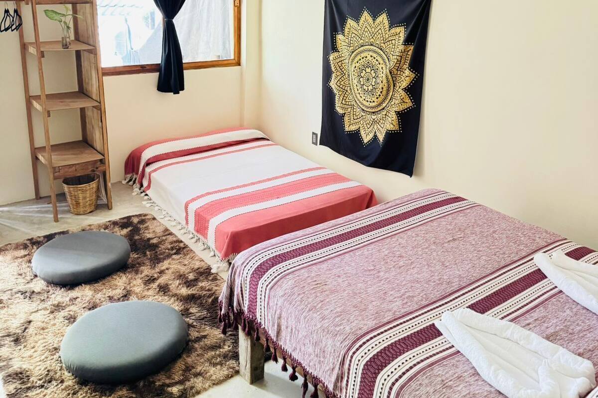 A room with two single beds, a ceiling fan, a hanging light fixture, a wooden shelf, a window with curtains, and a tapestry on the wall.