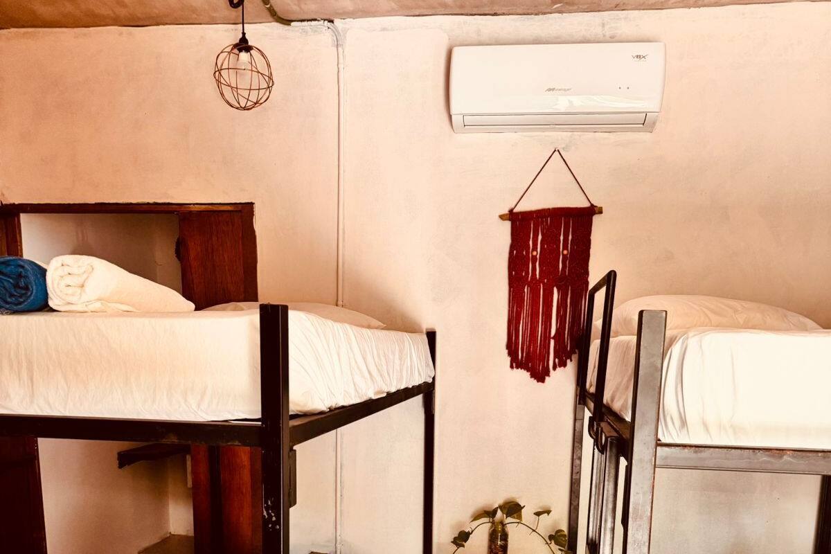 A dormitory room with bunk beds, a small wooden table, a hanging light fixture, and an air conditioning unit.