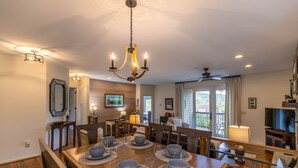 A dining area with a wooden table set for four, surrounded by chairs, a chandelier, and a television in the background.