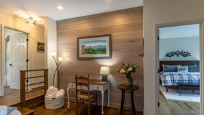 A bedroom with a bed, a bedside table with a lamp, a chair, and a desk with a chair. There is a painting on the wall and a door leading to another room.