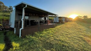 Un edificio moderno con un espacio al aire libre techado, un campo verde y una puesta de sol de fondo.