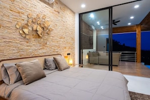Ein Schlafzimmer mit einer Steinsäule, einem Bett mit Kissen, einem Nachttisch und Blick auf einen Außenbalkon mit einem Stuhl.