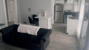 A modern living room with a black sofa, a small dining area with two chairs, and a kitchen with white cabinets and appliances.