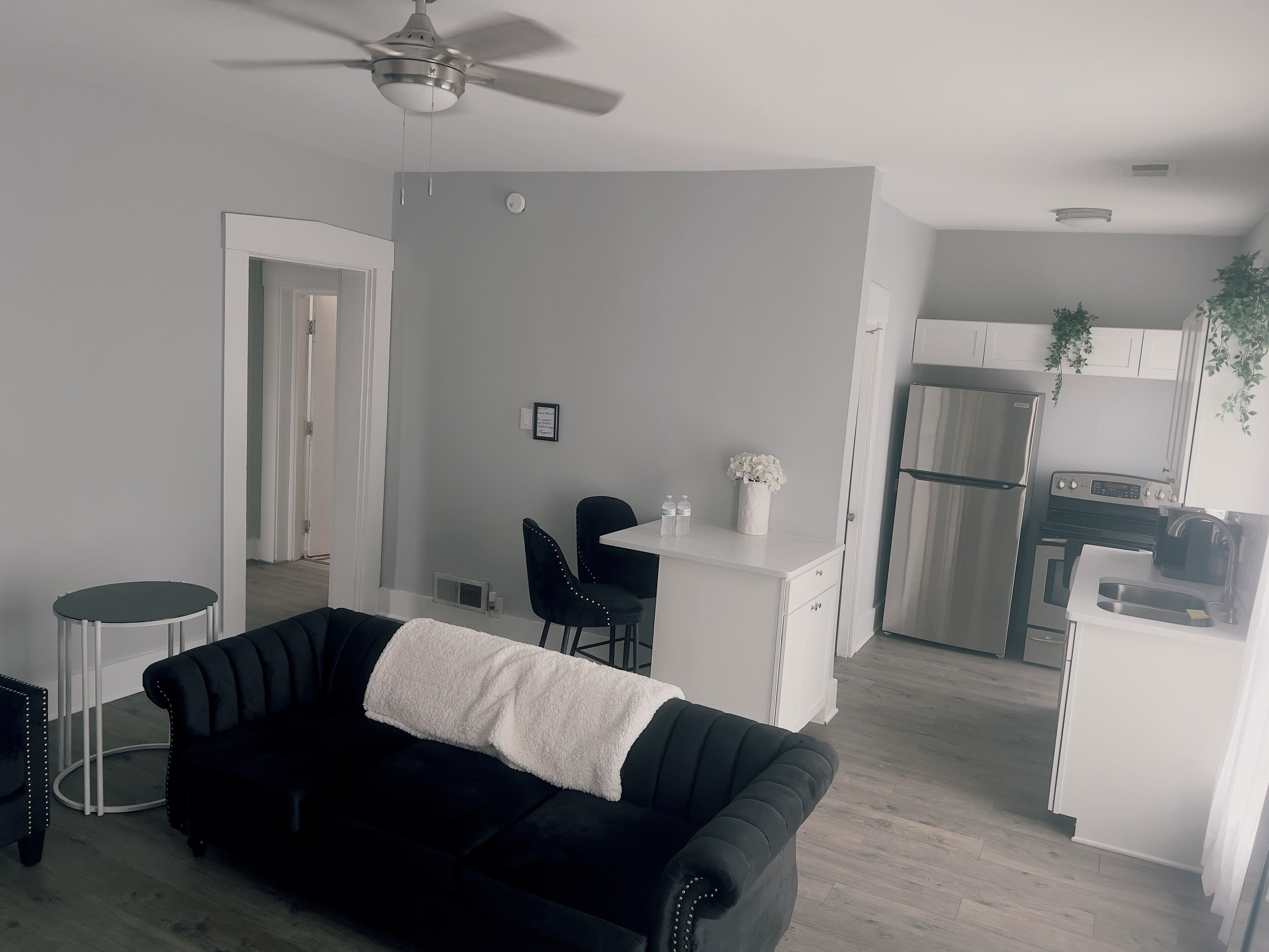 A modern living room with a black sofa, a small round table, and a ceiling fan. A kitchen area is visible in the background with white cabinets and appliances.