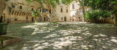 A courtyard with cobblestone paving, stone buildings with arched doorways, and trees providing shade.