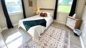 A bedroom with a bed, a nightstand, a bedside table, a rug, and a window.
