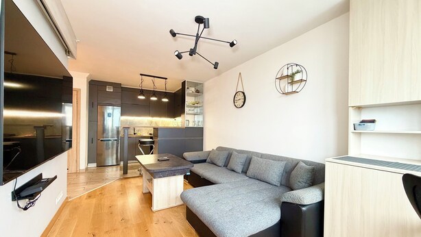 A modern kitchen with dark cabinets, a built-in oven, and a stainless steel refrigerator. There is a grey sectional sofa and a small table in the living area.