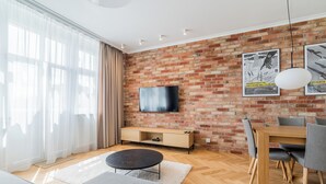 A modern living room with a brick wall, a flat-screen TV, a wooden dining table with chairs, and a grey sofa. - Kochanowskiego Luxury Apartment (Poznań)