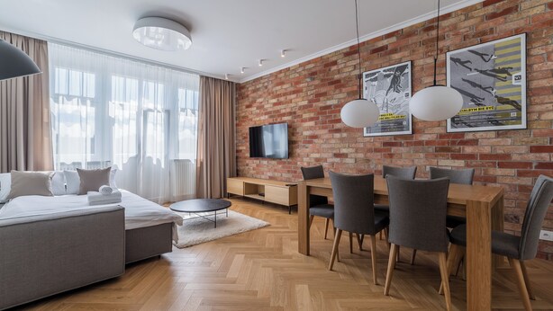A modern living room with a brick wall, a flat-screen TV, a dining area with wooden furniture, and a large window with sheer curtains. - Kochanowskiego Luxury Apartment (Poznań)