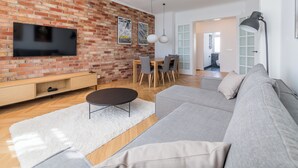 A modern living room with a brick wall, a flat-screen TV, a dining area with a table and chairs, and a gray sofa. - Kochanowskiego Luxury Apartment (Poznań)