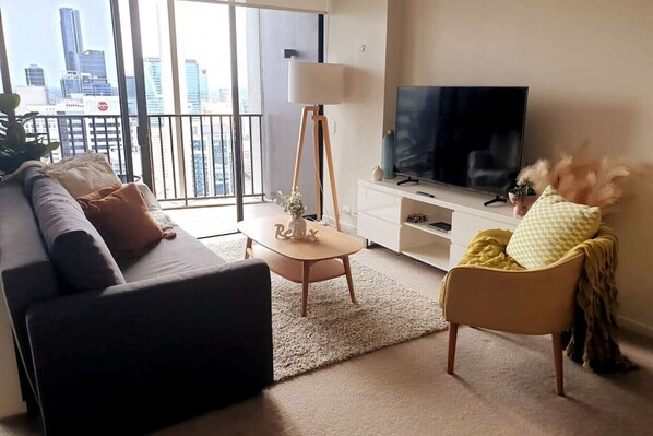 A modern living room with a sofa, a coffee table, a lamp, a TV on a stand, and a view of the city skyline through large windows. - InSpired Serenity on Queen St | 2 bed 2 Bath Apt | River Views Fast Wifi Carpark Gym Rooftop Pool (Brisbane City)