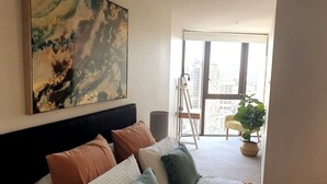 Une chambre avec une grande fenêtre, un lit avec des oreillers, un tableau et une plante.