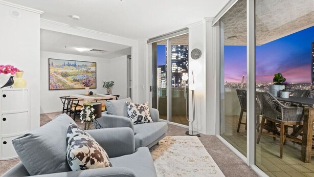A modern living room with a grey sofa, a dining table, and a cityscape view through large glass doors.
