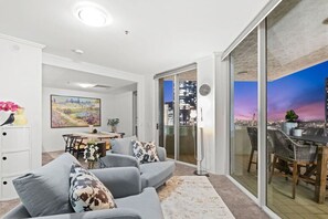 A modern living room with a grey sofa, a dining table, and a cityscape view through large glass doors.