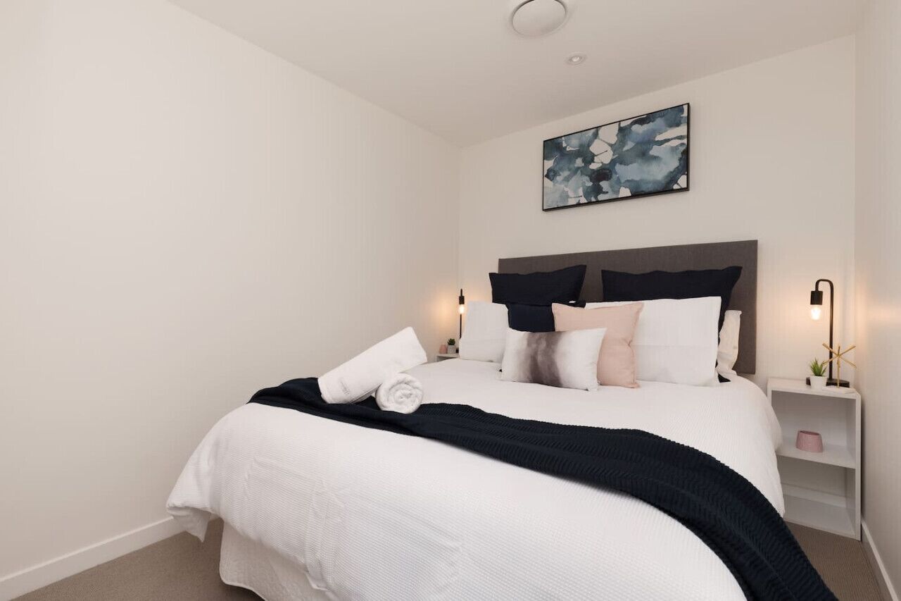 Een netjes opgemaakt bed met een zwart-wit dekbed, een donker hoofdbord en meerdere kussens. Een klein nachtkastje met een lamp en decoratie is zichtbaar.