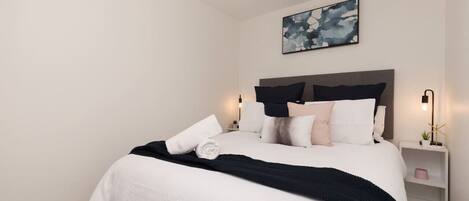 Een netjes opgemaakt bed met een zwart-wit dekbed, een donker hoofdbord en meerdere kussens. Een klein nachtkastje met een lamp en decoratie is zichtbaar.
