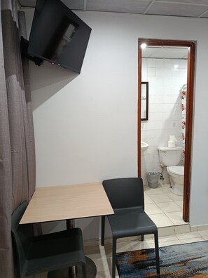 Une pièce avec un téléviseur fixé au mur, une table, deux chaises, une porte en bois donnant sur une salle de bain et un tapis à motifs sur le sol.
