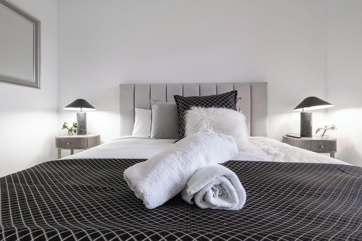 A neatly made bed with a rolled white towel, a grey headboard, and two bedside tables with lamps.