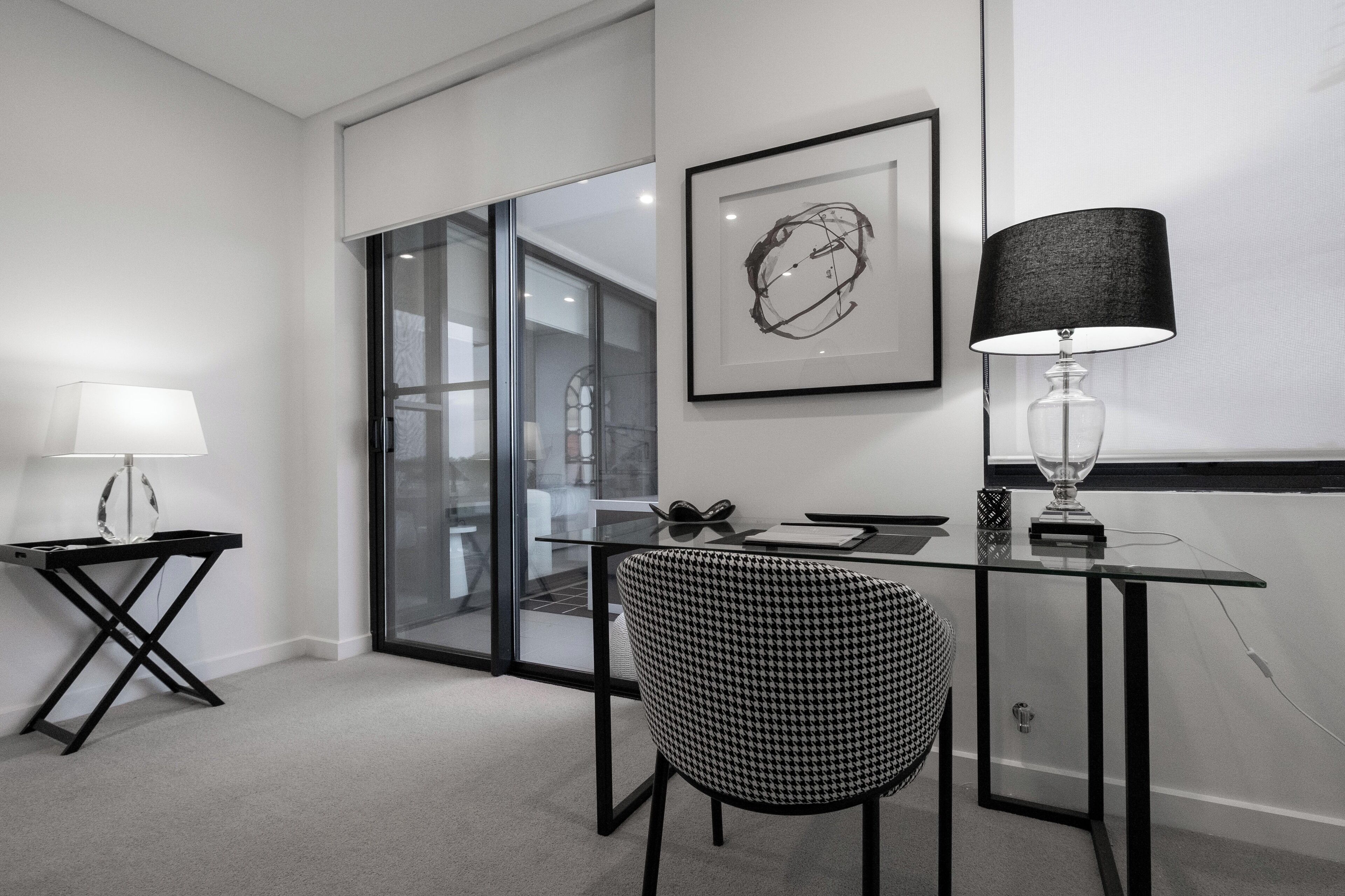 Une chambre d’hôtel moderne dotée d’une cloison en verre, d’un bureau avec une chaise, d’une lampe et d’une œuvre d’art encadrée au mur.