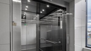 Un ascenseur moderne aux parois métalliques et à la porte en verre, doté d’une barre de maintien et d’un panneau indiquant la sortie de secours.