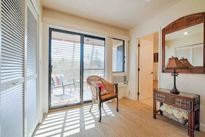 A room with a sliding glass door leading to a balcony, a wicker chair, a wooden console table with a lamp, and a large mirror.