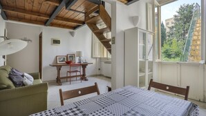 A room with a bed, a table, a chair, a sofa, a staircase, and a window with a view of greenery. - Angelico's Urban Loft (Firenze)