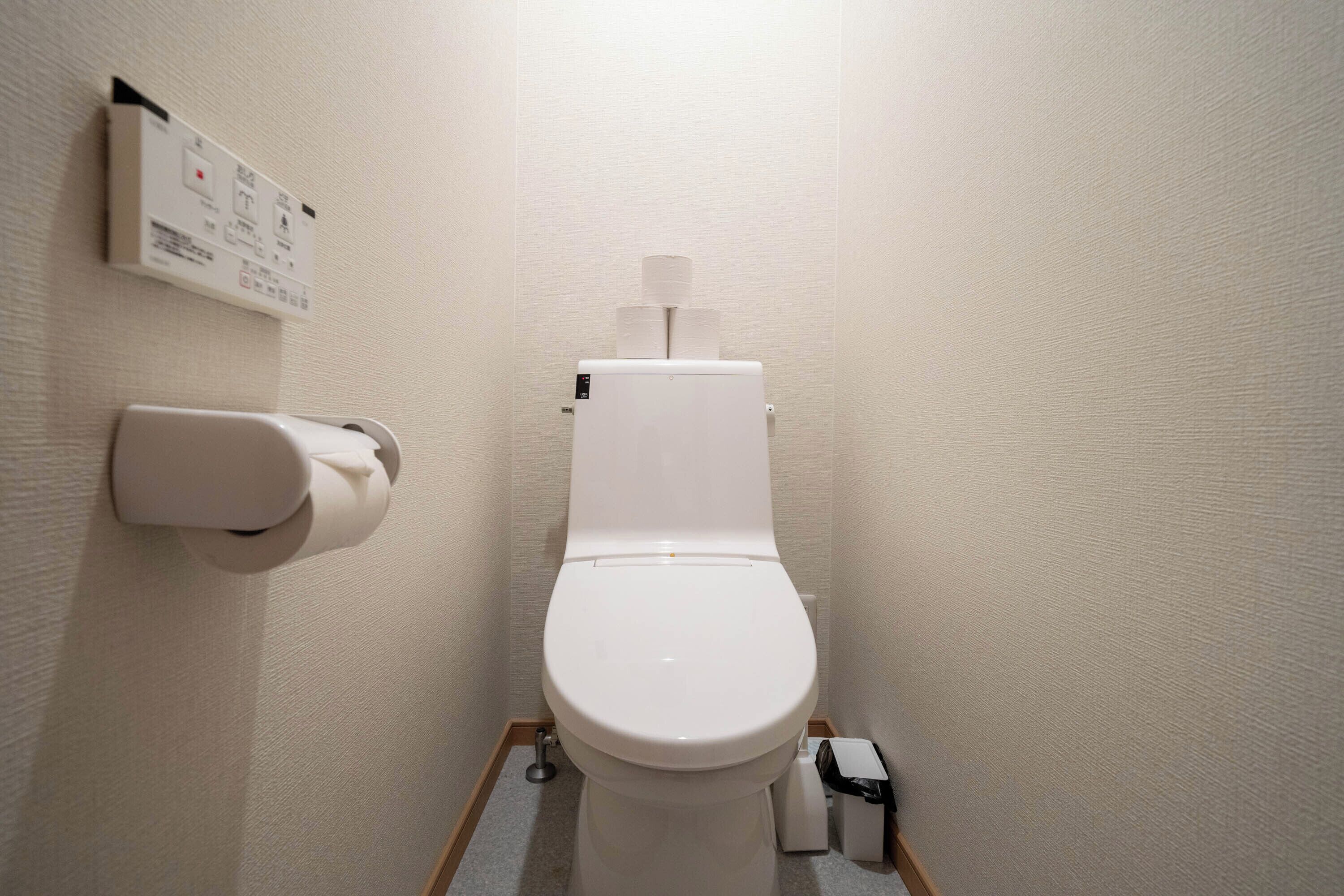 Inodoro blanco estándar con palanca de descarga, un dispensador de papel higiénico y un cubo de basura.