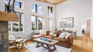 A spacious living room with a brown leather sofa, a wooden coffee table, and a stone fireplace. Large windows provide natural light and views of the surrounding area.