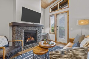 A cozy living room with a stone fireplace, a flat-screen TV mounted above it, and a comfortable seating area with a sofa and a chair.
