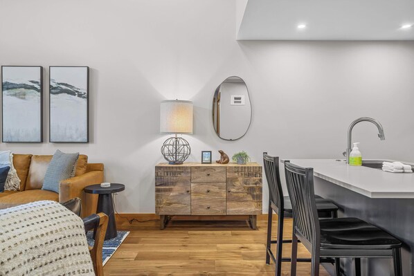 A modern, compact living space with a wooden floor, a sofa, a small side table, a cabinet with drawers, a round mirror, a lamp, a sink, and a chair.