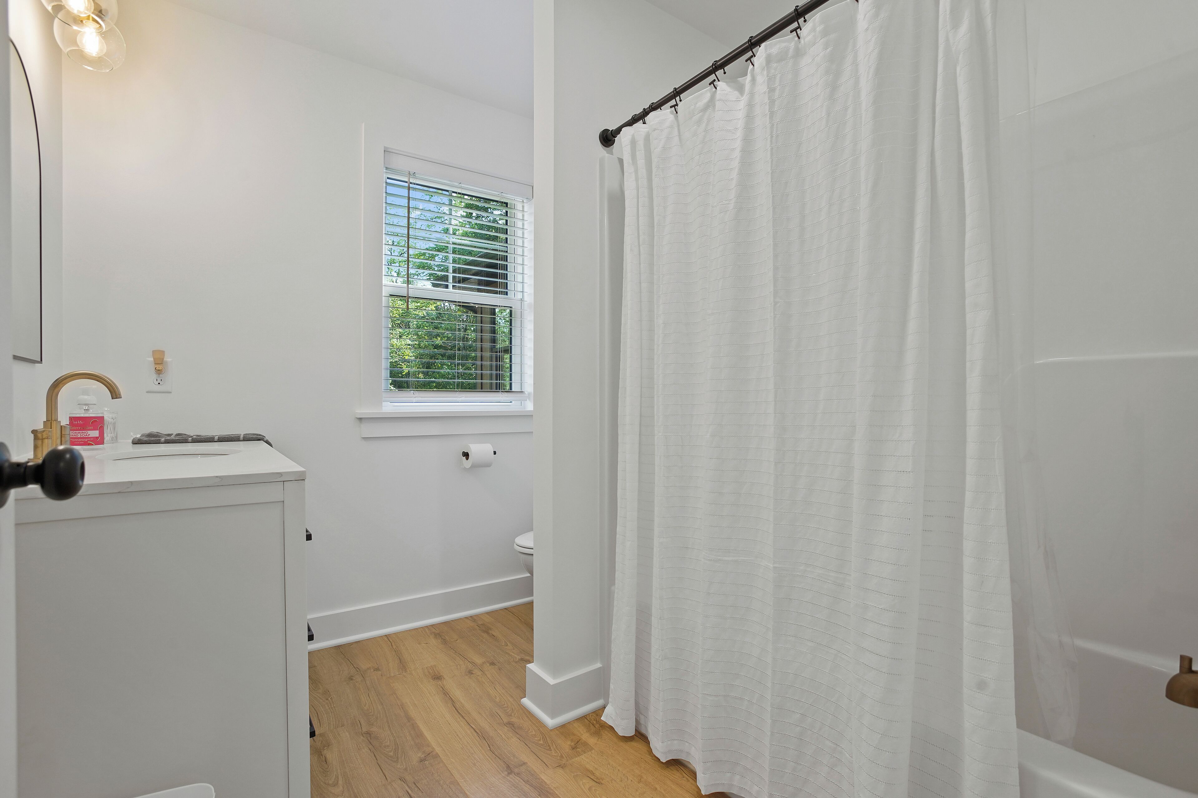 A bathroom with a toilet, a window with blinds, a shower curtain, and a sink with a soap dispenser.