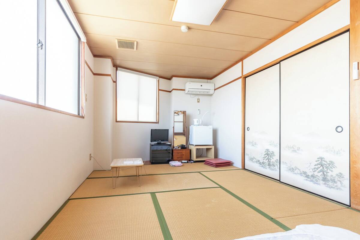 Une pièce de style japonais traditionnel, avec un sol recouvert de tatamis, un futon servant de lit, une télévision, un climatiseur et une porte coulissante ornée d’un paysage peint.