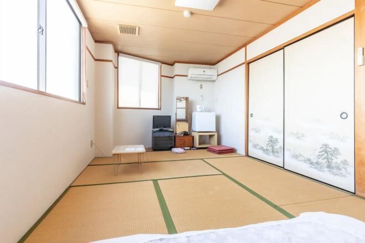 Een kamer met tatami vloerbedekking, een bed, een televisie en een schuifdeur met uitzicht op bomen.