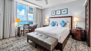 A hotel room with a large bed, a desk, a chair, a bench, a ceiling fan, and a view of the sky through the window. - Palm Beach Singer Island Resort & Spa Luxury Suites 2/2 Ocean View (Riviera Beach)