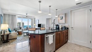 A modern hotel room with a kitchen area, a living area with a sofa and armchairs, and a large window offering a city view. - Palm Beach Singer Island Resort & Spa Luxury Suites 2/2 Ocean View (Riviera Beach)