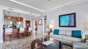 A modern living room with a sofa, coffee table, and bar stools. A kitchen with a microwave and refrigerator is visible in the background.