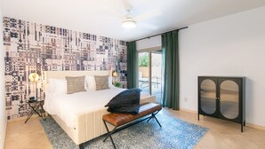 A bedroom with a large bed, a bench, a ceiling fan, a patterned wall, a cabinet, and a view of the outdoors. - Welcome to Casa Jubilant Cacti by HomeSlice Stays (Phoenix)