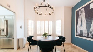 A dining area with a round table, six chairs, a chandelier, and a large window with blinds. - Welcome to Casa Jubilant Cacti by HomeSlice Stays (Phoenix)