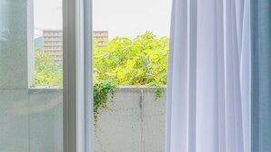 Blick durch ein Fenster mit weißen Vorhängen, einer Betonwand und Grünpflanzen draußen.