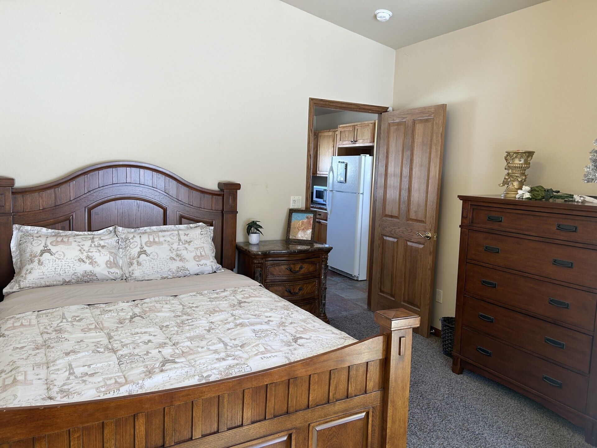 Une chambre à coucher avec un lit en bois, une table de chevet, une commode et une porte donnant sur la cuisine.