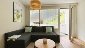 Uma sala de estar moderna com um sofá seccional, uma mesa de centro redonda de madeira e uma janela grande com persianas.