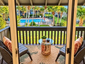 A balcony with wicker furniture, a glass table, and a view of a swimming pool and greenery.