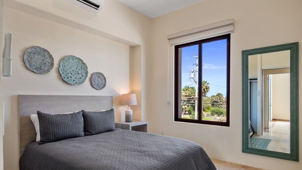 A bedroom with a large bed, a nightstand with a lamp, and a window with a view of palm trees. - Tormenta 202 3BR Second Floor San Jose del Cabo (San José del Cabo)