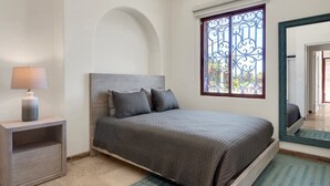 A bedroom with a bed, a nightstand, a lamp, a window with decorative iron bars, and a mirror. - Tormenta 101 3BR Ground Floor San Jose del Cabo (San José del Cabo)