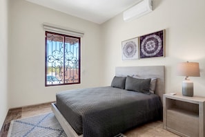 A bedroom with a bed, a nightstand, a lamp, two wall art pieces, a window with a metal gate, and a ceiling fan. - Tormenta 101 3BR Ground Floor San Jose del Cabo (San José del Cabo)